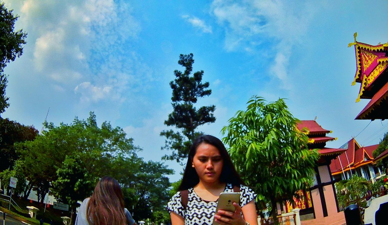 tree, sky, person, looking at camera, lifestyles, building exterior, leisure activity, young adult, architecture, casual clothing, built structure, portrait, smiling, low angle view, cloud - sky, front view, cloud, day