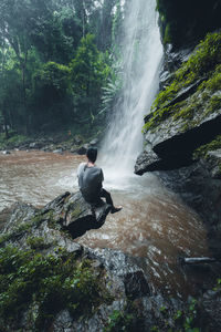 Rear view of man sitting on rock against waterfall in forest