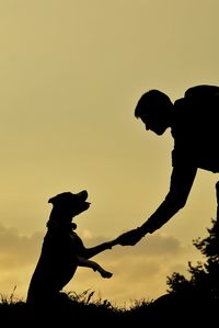 Silhouette man giving handshake to dog against sky during sunset