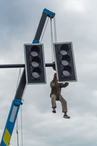 Low angle view of man hanging on road signal against sky