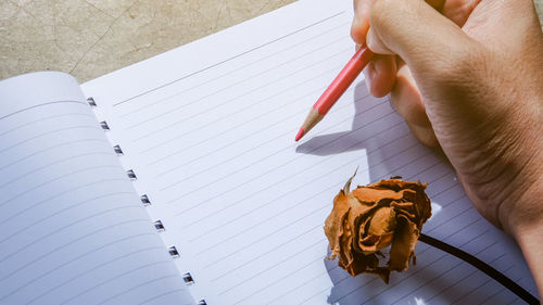 Close-up of human hand holding pencil over book by wilted flower on table