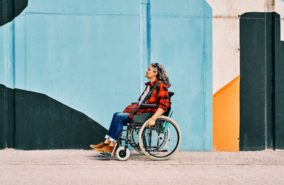 Full body side view of independent handicapped mature female riding wheelchair near painted wall on street