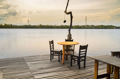 Chairs and table by lake against sky during sunset