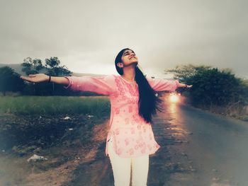 Young woman with arms raised standing on field against sky