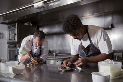 Concentrated black male chef cooking dish near colleague squeezing cream out of pastry bag on appetizer in kitchen of restaurant
