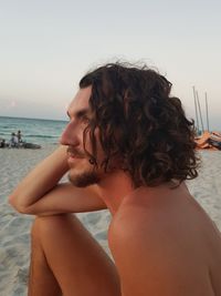 Side view of shirtless man sitting at beach against sky