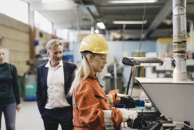 Female industrial worker in hardhat working on machinery while managers in background at factory