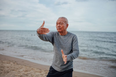 Senior man exercising while standing at beach against cloudy sky during sunset
