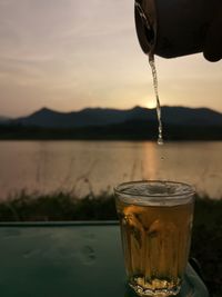 Close-up of beer glass against lake during sunset