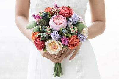 Midsection of bride holding bouquet while standing against white background