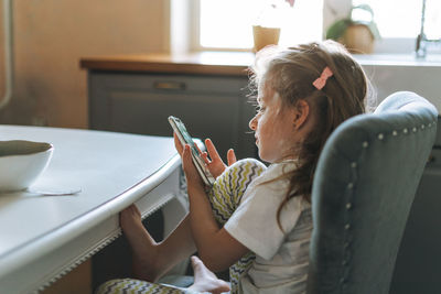Little child beautiful girl with long hair in home clothes using mobile at dinning table in kitchen 