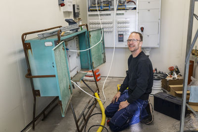 Smiling male electrician kneeling by electric panel in industry