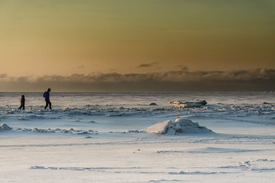 People walking in frozen sea against sky during sunset