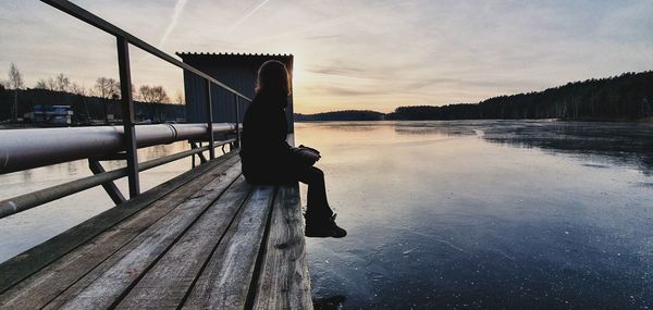 Man sitting on pier over lake against sky during sunset