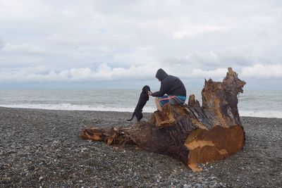 Man sitting on driftwood at beach against sky