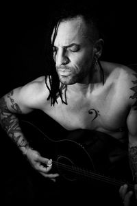 Man playing guitar and singing shirtless. dark background