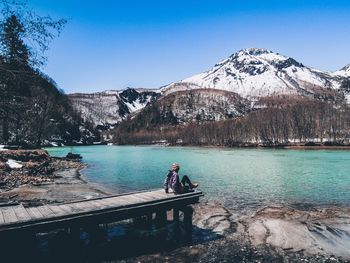 Woman sitting on jetty by river against snowcapped mountain