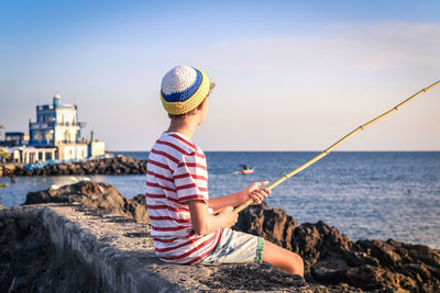 Boy fishing while sitting on rock at beach against sky