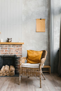 A wicker armchair with a yellow cushion near a brick fireplace with firewood