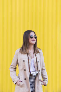 Fashionable young woman wearing sunglasses and overcoat while standing against yellow wall