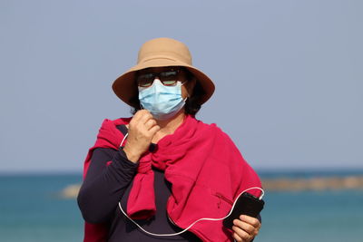 Portrait of woman wearing mask and hat standing against sea against clear sky