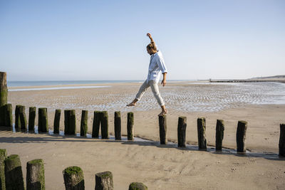 Young man balancing while walking on wooden posts at beach against clear sky