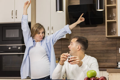 Surprised man looks at dancing pregnant female in kitchen, male has tea. woman's hormones change