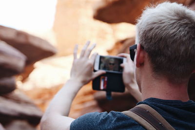 Rear view of man by rock formation taking selfie from mobile phone