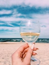 Cropped image of woman holding wineglass at beach against sky