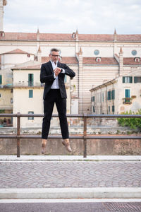 Full length of man jumping against building while checking time in watch