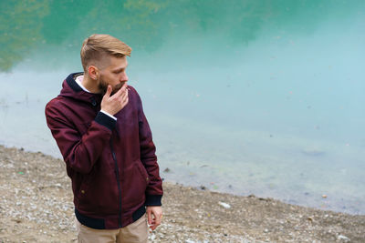 Young bearded man stood alone by lake and looked aside thoughtfully smoking