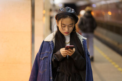 Korean young female texting online at subway metro station hold smartphone, read sms or book tickets