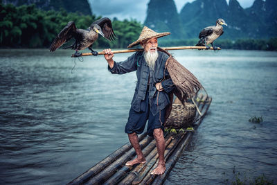 Birds perching on stick held by man on wooden raft in lake
