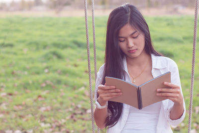 Young woman reading book while sitting on swing in park