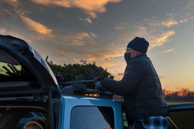 Man wearing mask standing by car against sky