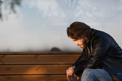 Pensive serious young bearded man wear leather jacket sitting and thinking outdoors