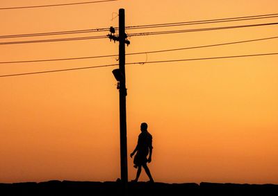 Silhouette man walking by electricity pylon against clear sky during sunset