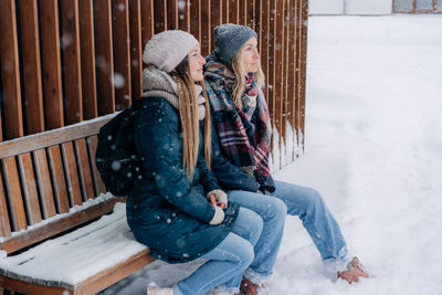 Two girlfriends sit on a snow-covered bench in the winter on the street and enjoy the weather.