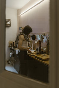 Young man wearing undershirt preparing food while standing in kitchen at home seen through door