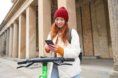 Young woman using mobile phone while standing against wall