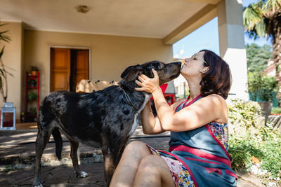 Grey dog licking it's female owner who is sitting in front of the house