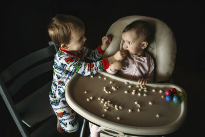 Brother feeding baby food to sister sitting on high chair in darkroom at home