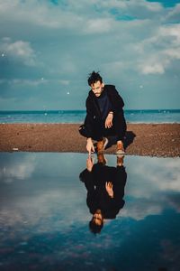 Young man in crouching by sea on beach against sky