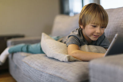 Smiling boy using tablet computer while lying on sofa at home