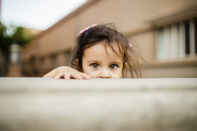 Portrait of cute girl hiding behind retaining wall against house