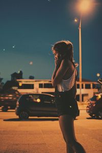 Side view of woman talking on mobile phone while standing on illuminated street at night