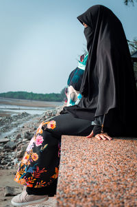 Side view of woman in burka sitting on retaining wall against sky