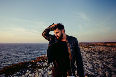 Mid adult man with hand in hair standing by sea against sky during sunset