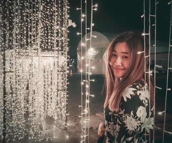 Portrait of smiling woman standing by illuminated string lights at night