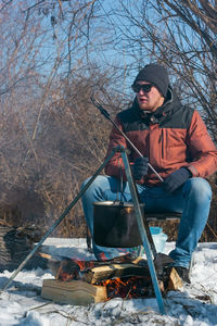 Man by campfire, pot of soot over bonfire hanging on tripod, winter outdoor cooking at campsite